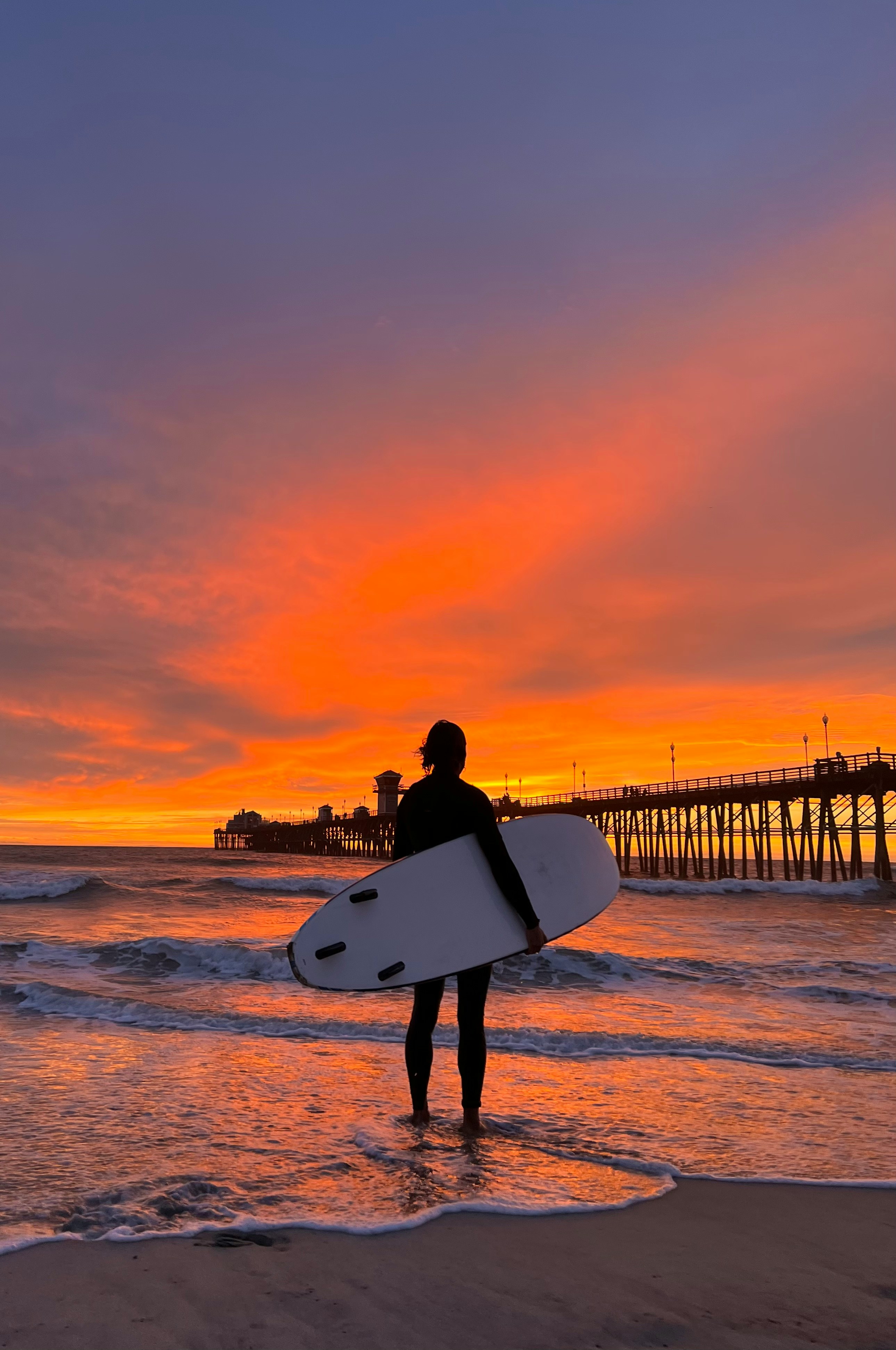 A surfer holding a board at sunset beside the Oceanside Pier